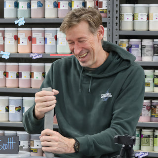 A potter laughing hard while gripping a long gray flat object in front of shelves of glaze jars at The Clay Hole in Draper, Utah