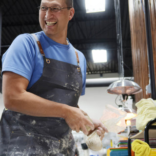 A laughing potter in a clay-dusted apron holds a plastic-wrapped bundle of clay and looks off to the side at The Clay Hole in Draper, Utah