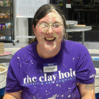 A laughing person in a purple t-shirt with wet clay smeared across the face and splatters on the shirt at The Clay Hole in Draper, Utah