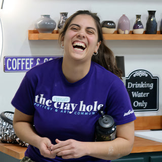 A laughing potter in a purple t-shirt with hands clasped, with a wall shelf of finished vases behind at The Clay Hole in Draper, Utah
