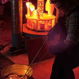 A potter in a headlamp holds long tongs by a glowing orange platform holding cups, vases and a pierced cylinder at The Clay Hole in Draper, Utah