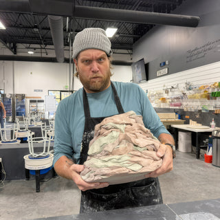 A potter in a gray beanie holds a large chunk of layered pink and gray clay with a stern look at The Clay Hole in Draper, Utah