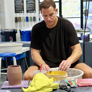A potter works over a bucket at a table, with a tall mauve clay cup in the foreground at The Clay Hole in Draper, Utah