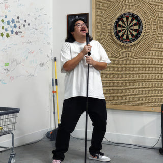 A member performs into a microphone on a stand in front of a dartboard and a wall covered in signatures at The Clay Hole in Draper, Utah