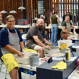Two smiling potters sit at pottery wheels shaping small clay forms, with a third at a wheel behind them at The Clay Hole in Draper, Utah