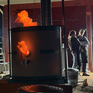 A mesh kiln glows orange with flames from the top and a side hole while two people stand behind it at The Clay Hole in Draper, Utah