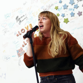 A person in a striped rust and green sweater talking into a microphone in front of a signature wall at The Clay Hole in Draper, Utah