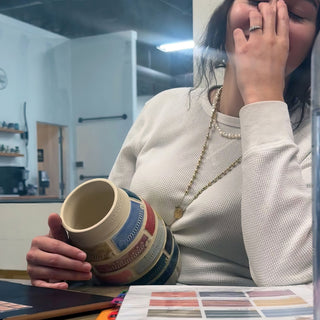 A person with a hand over their face holds a cream mug wrapped in blue and red patterned bands at The Clay Hole in Draper, Utah
