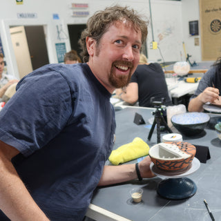 A smiling potter at a table beside a bowl painted with orange and black patterns sitting on a turntable at The Clay Hole in Draper, Utah