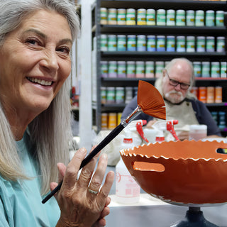 A smiling potter holding an orange-bristled fan brush beside an orange bowl with a scalloped rim and cut-out handles at The Clay Hole in Draper, Utah