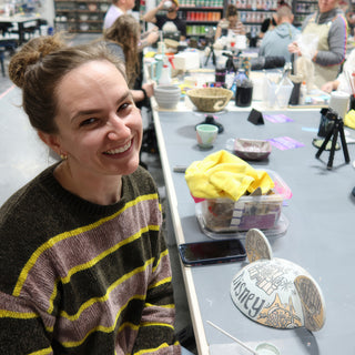 A smiling potter in a striped sweater at a long worktable with a painted half-round piece in front at The Clay Hole in Draper, Utah