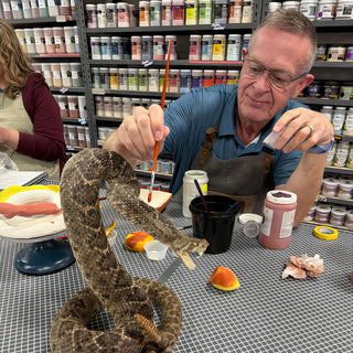 A potter paints with a thin brush at a gridded worktable beside a coiled snake with a brown patterned body at The Clay Hole in Draper, Utah
