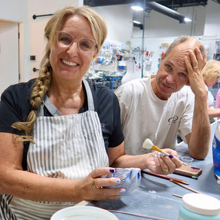Two potters sit at a table, one holding a small bowl with blue and white wavy patterns and a paintbrush at The Clay Hole in Draper, Utah