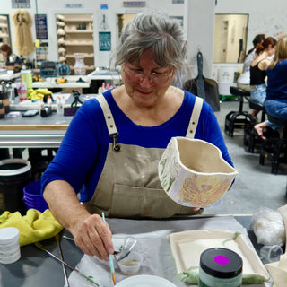 A potter holds up an irregular cream cup painted with leaves and a yellow winged figure, brush in hand at The Clay Hole in Draper, Utah