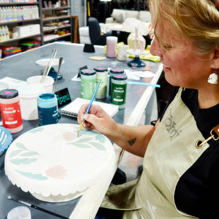 A potter paints pink flowers and green leaves with a thin brush on a white plate with a scalloped edge at The Clay Hole in Draper, Utah
