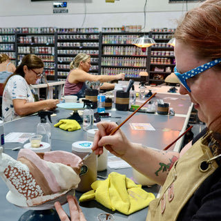 A potter in blue polka dot glasses paints a shallow bowl striped in pink, brown and cream with white dots at The Clay Hole in Draper, Utah