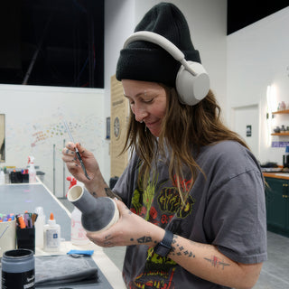 A potter in a beanie and headphones paints a small white bottle-shaped vase with a thin brush at The Clay Hole in Draper, Utah