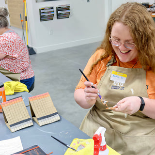 A laughing potter in an orange shirt painting a small clay piece with a thin brush beside two grid-patterned pieces at The Clay Hole in Draper, Utah