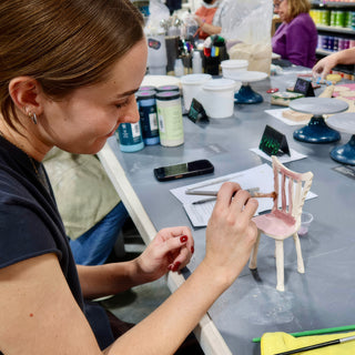 A potter painting pink onto the seat and back of a small white ceramic chair with a thin brush at The Clay Hole in Draper, Utah