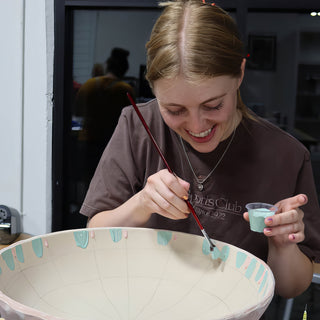 A potter smiles while brushing teal drips onto the rim of a large bowl with pencil guide lines at The Clay Hole in Draper, Utah