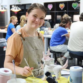 A potter in a tan apron smiling while brushing a white sculpted figure with pink-tinted wings and horns at The Clay Hole in Draper, Utah