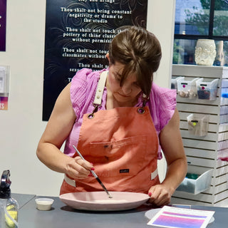 A potter in a coral apron holds a thin brush over a wide pale pink plate at a work table at The Clay Hole in Draper, Utah