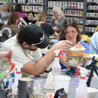 Two potters smiling over a pale bowl on a turntable, with a tiered white clay cone and spray bottles at The Clay Hole in Draper, Utah