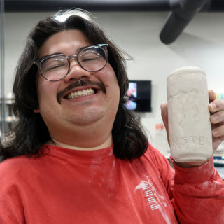 A smiling potter in a red shirt holds up a pale cylinder shaped like a tall can with raised lettering at The Clay Hole in Draper, Utah