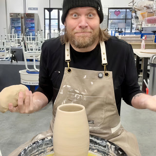 A wide-eyed potter in a tan apron holds a lump of clay beside a tall pale cylinder on a wheel at The Clay Hole in Draper, Utah