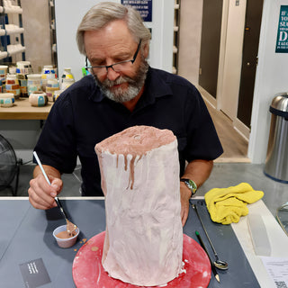 A potter with glasses brushing brown color onto a tall pale clay cylinder on a red board at The Clay Hole in Draper, Utah