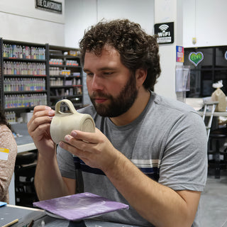 A potter with a beard examines a pale clay mug with a looped handle, held in both hands at The Clay Hole in Draper, Utah
