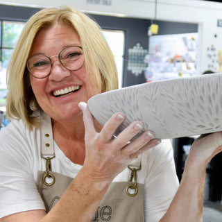 A smiling potter in round glasses holds up a pale gray bowl marked with feathery leaf-like lines at The Clay Hole in Draper, Utah
