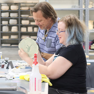 Two potters at a worktable, one holding up a pale green scalloped plate with a darker green figure at The Clay Hole in Draper, Utah