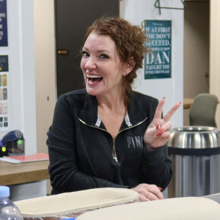 A laughing potter in a black zip jacket flashes a peace sign behind two oval clay slabs on a table at The Clay Hole in Draper, Utah