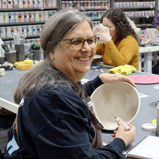 A smiling potter in a navy sweatshirt holding a pale shallow bowl and a small pointed tool at a worktable at The Clay Hole in Draper, Utah