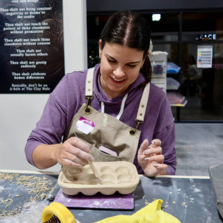 A smiling potter using a small tool to work inside a pale tray with six oval compartments at The Clay Hole in Draper, Utah