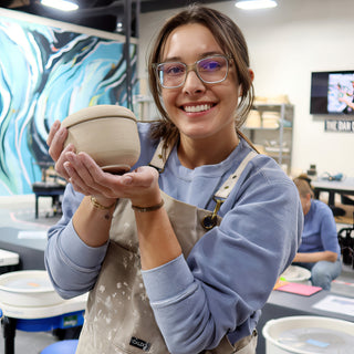 A smiling potter in glasses and a tan apron cradling a small pale unglazed bowl with a ridged rim at The Clay Hole in Draper, Utah