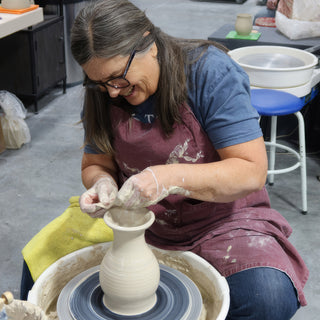 A potter in a maroon apron laughing while shaping the neck of a pale ridged vase on a pottery wheel at The Clay Hole in Draper, Utah