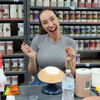 A potter laughing with a thin brush and a tiny orange cup in front of a peach-colored bowl at The Clay Hole in Draper, Utah