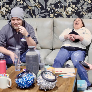 Two people laugh on a couch behind a table with a tall dark vase and two bowls covered in beads at The Clay Hole in Draper, Utah