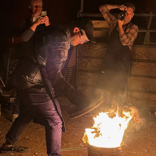 A person in a cap and apron bends toward a burning metal can while two people behind take photos at The Clay Hole in Draper, Utah