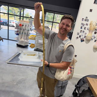 A smiling person holds up a very long strip of yellow tickets with one arm, a tote on their shoulder at The Clay Hole in Draper, Utah