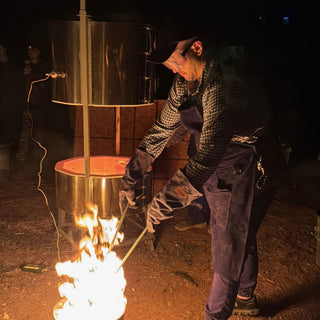 A person in a cap, goggles and checkered shirt holds two long rods toward flames beneath a steel cylinder at The Clay Hole in Draper, Utah