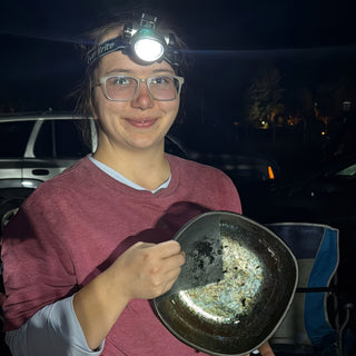 A smiling person in glasses and a headlamp holds a shallow metal bowl with dark flecks inside, outdoors at night, with The Clay Hole, a pottery studio in Draper, Utah