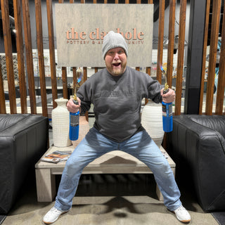 A person in a beanie sits legs apart holding two lit blue torches beside white ribbed vases at The Clay Hole in Draper, Utah