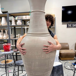 A person peeks out from behind a tall taupe vase and wraps both arms around its wide body at The Clay Hole in Draper, Utah