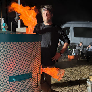 A person in a headlamp stands beside a teal mesh kiln with flames coming out of the top and side at The Clay Hole in Draper, Utah