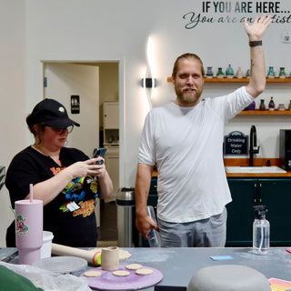 A person waves at the camera in the studio while another in a cap looks at a phone at The Clay Hole in Draper, Utah