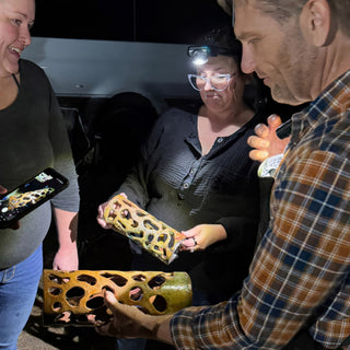 Several people gather around a cylinder with organic cutouts in amber and green, one taking a phone photo of it at The Clay Hole in Draper, Utah