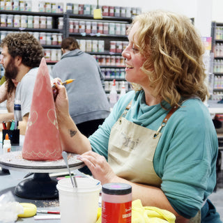 A potter in a teal sweatshirt decorates a tall pink clay cone with scalloped lines on a turntable at The Clay Hole in Draper, Utah